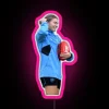 AFL player Steph Catley poses with an Australian Rules football and a vibrant neon sign in pink hues.