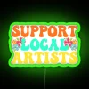 Vibrant RGB neon sign encouraging support for local musicians, with green and blue hues in a futuristic music-themed display.