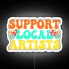 Neon sign promoting support for local artists with colorful letters on a dark background.
