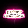 Neon sign reading 'Support Your Local Animal Shelter' in pink letters against a dark background.