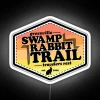 A neon-lit sign in a vibrant, rainbow color scheme advertises "Swamp Rabbit Trail" along a scenic outdoor path.