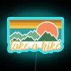 Neon sign with colorful, hand-painted letters reading 'Take A Hike' in bold, blue font on a dark background.